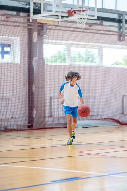 Dark-haired boy in sportswear running after the ball in a gym - Fotó, kép