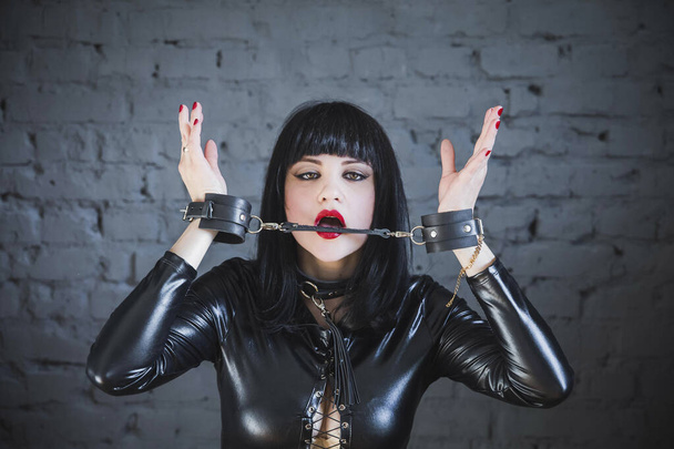 beautiful brunette in a black leather suit and collar gnaws at handcuffs - Photo, Image