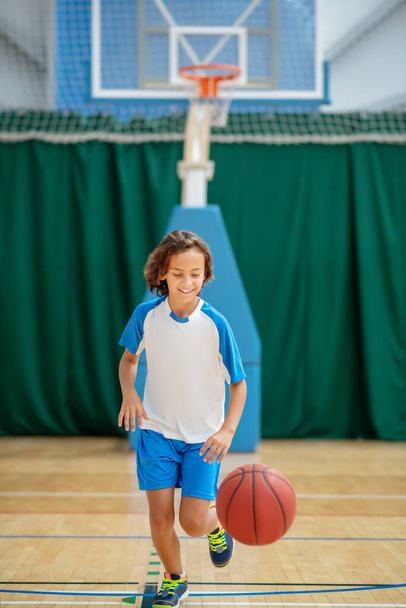 Dark-haired boy in sportswear training his basketball skills in a gym - Фото, изображение
