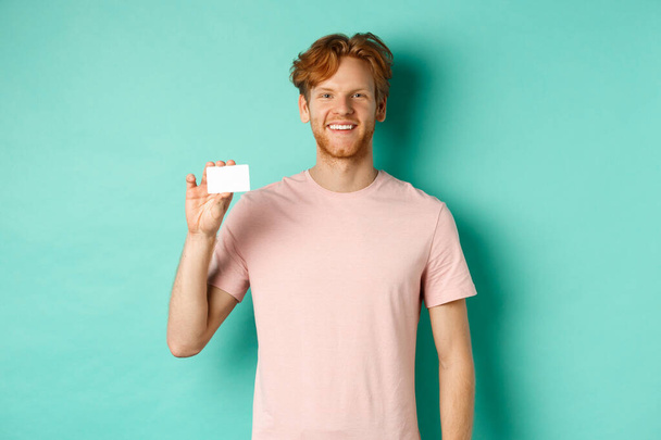 Hezký mladý muž usmívá a ukazuje plastové kreditní karty, stojící v tričku na tyrkysovém pozadí - Fotografie, Obrázek