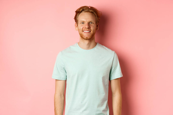 Happy redhead man in t-shirt looking at camera and smiling, standing over pink background - Photo, Image