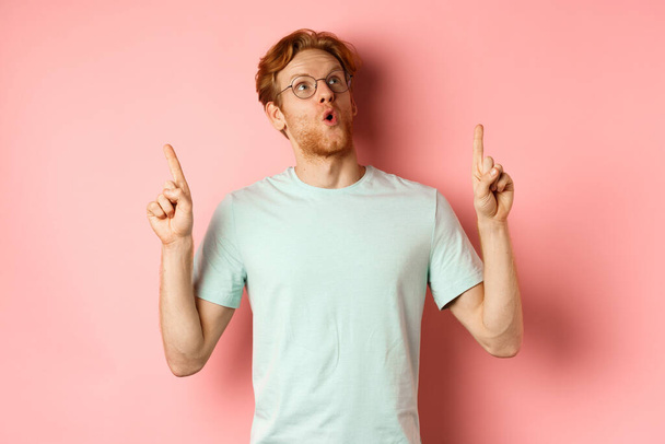 Wow check this out. Surprised redhead guy in glasses looking and pointing up, gasping amazed, standing over pink background - Foto, imagen
