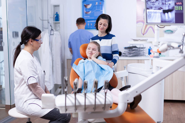Petite fille dans un cabinet de dentiste pédiatrique touchant le visage avec une expression douloureuse - Photo, image
