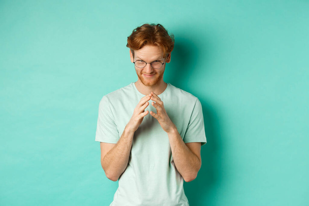 Devious redhead man in glasses and t-shirt pitching an idea, steeple fingers and look from under forehead with sly and smug smile, standing over mint background - Photo, Image