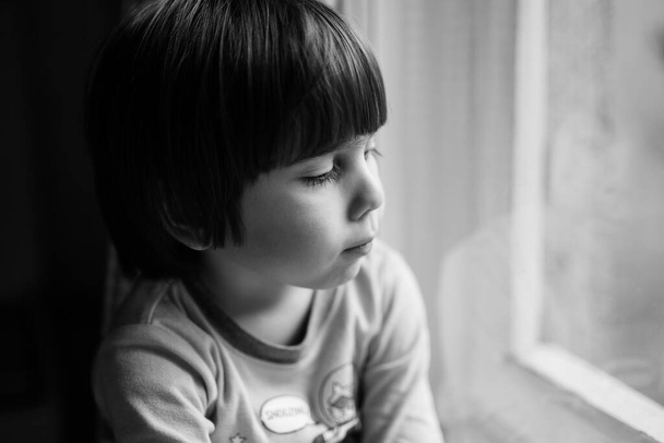 Un niño preescolar mirando por la ventana de su casa. Auténtico retrato en blanco y negro de un lindo niño en el interior. Enfoque franco con enfoque selectivo. - Foto, imagen