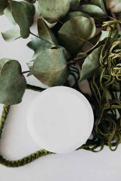 witte ronde cosmetische pot op gedroogde eucalyptusstengels en bladeren in groene snaarzak. redelijke consumptie van dingen, welzijnsfilosofie, selectieve focus - Foto, afbeelding