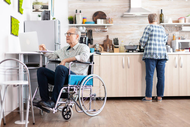 Elderly disabled person working on laptop in kitchen - Photo, Image