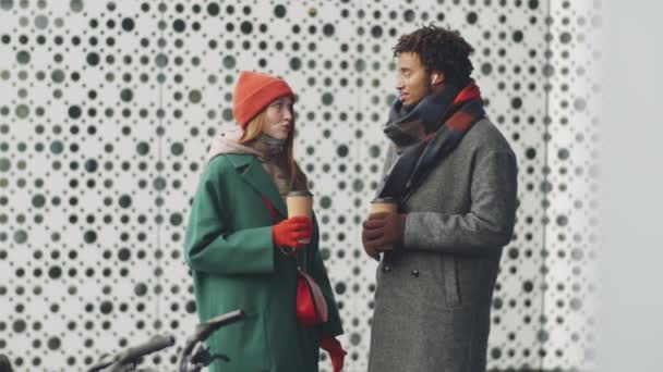 Mittlere Gesamtaufnahme eines jungen schwarzen Mannes und einer kaukasischen Frau, die auf der Straße neben E-Scootern stehen, Kaffeetassen halten und an Wintertagen im Freien diskutieren - Filmmaterial, Video