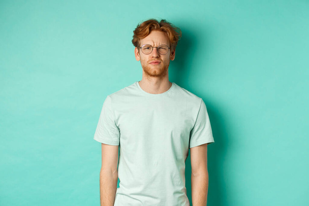 Confused young redhead man in glasses staring at camera puzzled, squinting perplexed, cant understand something, standing over mint background - Photo, Image