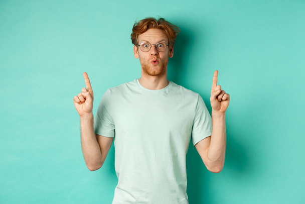 Beeindruckter rothaariger Mann in Brille und T-Shirt, Promo-Angebot checkend, Finger nach oben zeigend, staunend in die Kamera starrend, vor türkisfarbenem Hintergrund stehend - Foto, Bild
