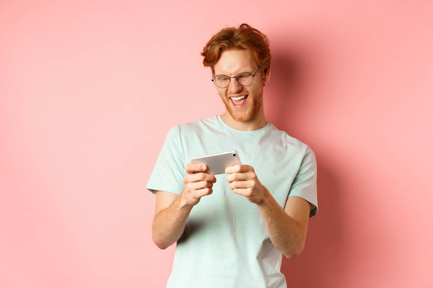 Happy young man with red messy haircut, wearing glasses, playing video game on smartphone and having fun, looking at mobile screen, standing over pink background - Fotografie, Obrázek