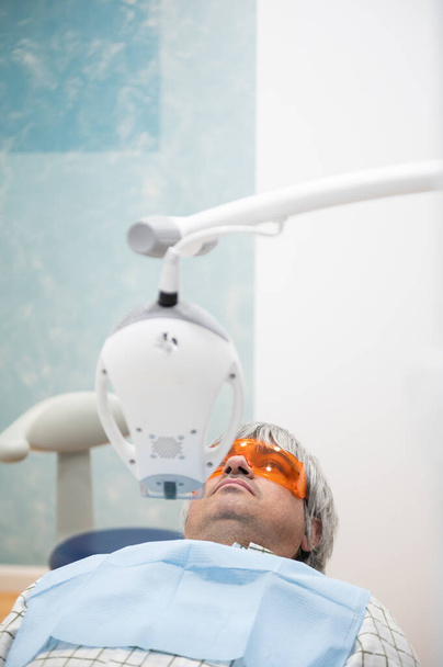 Mature man comfortably seated in dentist's chair with protective goggles ready to have his teeth whitened.Copy space.Dental and health care concept - Fotografie, Obrázek
