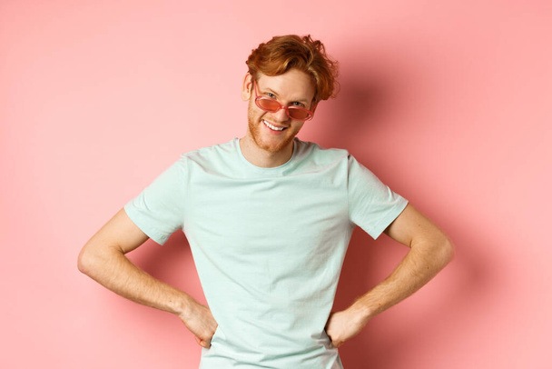 Concepto de turismo y vacaciones. Hombre barbudo divertido con el pelo rojo, con gafas, sonriendo y mirando desde debajo de la frente con las manos en las caderas, de pie sobre el fondo rosa - Foto, imagen
