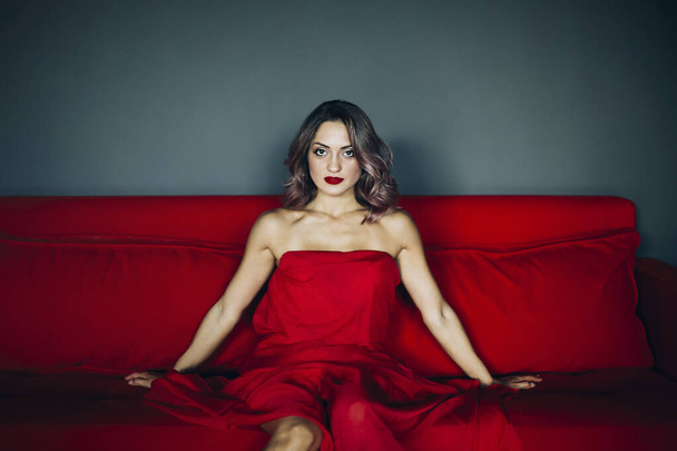 Atractiva sensual mujer joven con hombros desnudos con el pelo ondulado con vestido rojo sentado en el sofá rojo y mirando a la cámara - Foto, imagen