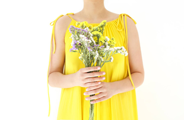 Mujer irreconocible en vestido amarillo escondido detrás de ramo de flores silvestres en el día de verano sobre fondo blanco - Foto, Imagen