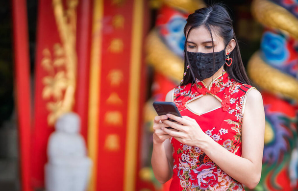 Asian young woman wearing red traditional Chinese cheongsam decoration and wear protective mask and smartphone for Chinese New Year Festival at shrine,Prevention of the spread of COVID-19 virus - Фото, изображение