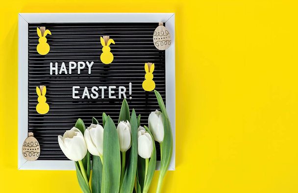 HAPPY EASTER is written on letter board, tulips flowers, and decorative wooden eggs on yellow background. Copy space, top view. Flat lay of Easter celebration concept.  - Photo, image