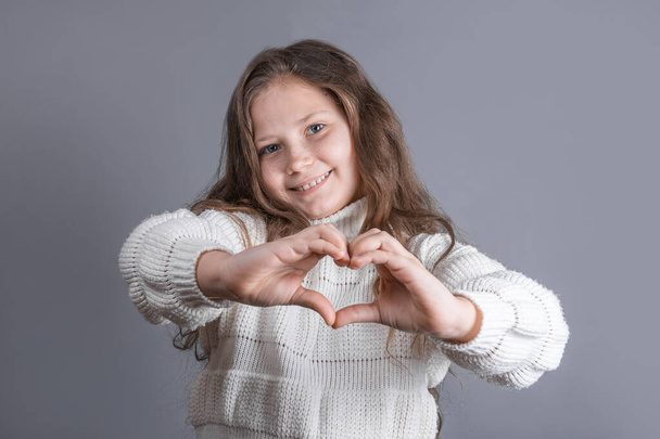 Portrait of a young attractive little girl with blond long flowing hair in a white sweater smiling shows heart hands on a gray studio background. Place for text. Copy space. - Фото, изображение