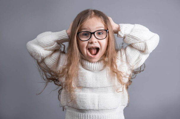 Portrait of surprised young attractive little girl with blond hair in sweater cheerful cheery straight-haired girls omg facial expression shock gesture on a gray studio background.Unexpected shocking. - Foto, Bild