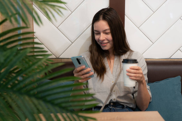 young woman is sitting in a cafe, drinking coffee in a cardboard glass - Фото, зображення