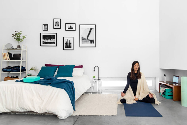 Junge Frau praktiziert Yoga und Meditation in Lotus-Pose im Schlafzimmer mit minimalistischem Interieur - Foto, Bild