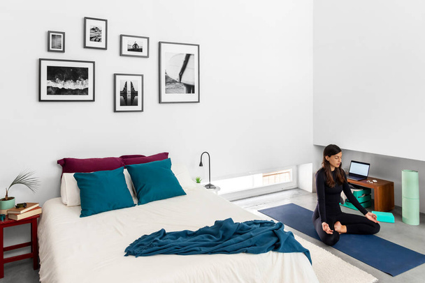 Jonge vrouw beoefenen van yoga en meditatie in lotus poseren in de slaapkamer met minimalistische stijl interieur - Foto, afbeelding