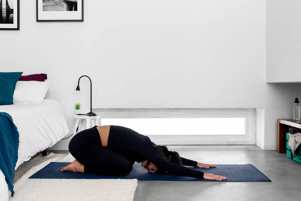 Joven mujer delgada practicando yoga en posición infantil extendida en el moderno dormitorio minimalista en casa - Foto, imagen