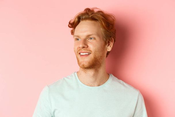 Portrait de gars caucasien attrayant avec des cheveux rouges désordonnés et la barbe, tourner la tête et regarder à gauche avec sourire heureux, debout sur fond rose - Photo, image