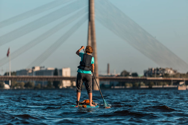 женщина грести с SUP встать весла доски вдоль реки, на заднем плане кабельный мост - Фото, изображение