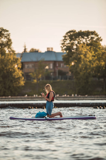 Une jeune femme blonde au téléphone sur un SUP se lève planche à pagaie dans la rivière en été, heure d'or - Photo, image