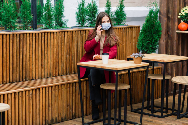 Close up portrait of a caucasian female wearing a medical mask and standing in the street against background of a cafe - Photo, image