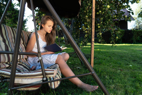 Ένα έφηβο κορίτσι διαβάζει ένα βιβλίο που κάθεται στην κούνια του κήπου στον καλοκαιρινό κήπο κάτω από μια μηλιά - Φωτογραφία, εικόνα
