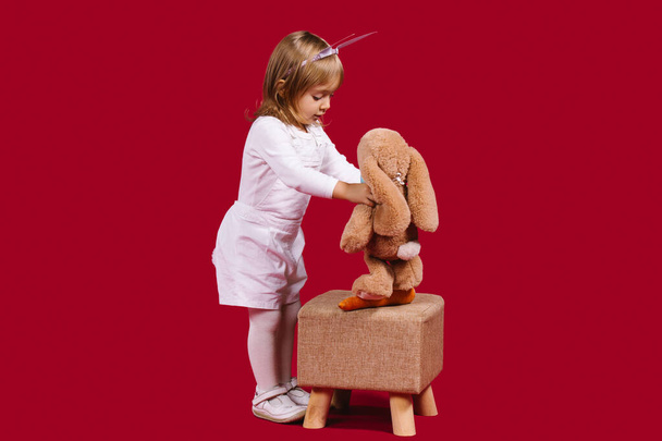 Caucasian kindergarten girl is played with a soft toy on a red background. The girl sits a soft bunny on a stool. Blank space for your advertisement. - Foto, afbeelding