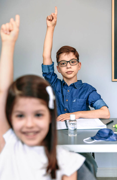 Μαθητές με μάσκα στο τραπέζι σηκώνουν τα χέρια στο σχολείο - Φωτογραφία, εικόνα