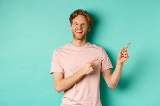 Beau modèle masculin avec des cheveux rouges désordonnés montrant la publicité sur l'espace de copie, pointant vers le coin supérieur droit et souriant heureux, debout sur fond turquoise - Photo, image