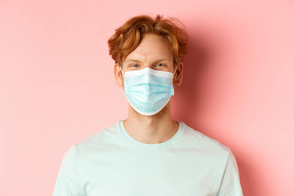 Covid-19 and pandemic concept. Close up of redhead man in medical mask looking at camera, standing over pink background - Fotografie, Obrázek