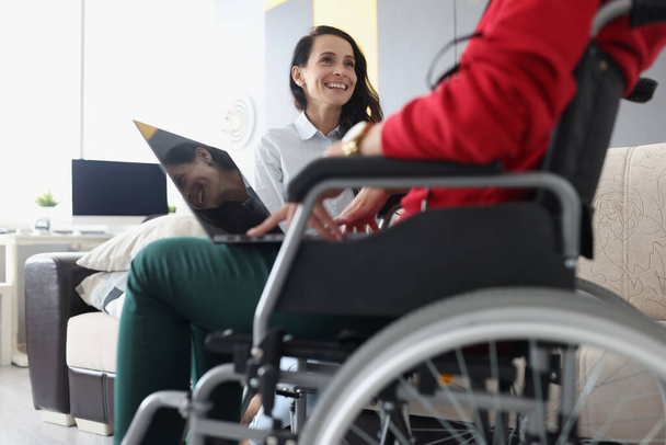 Χαμογελαστή γυναίκα επικοινωνεί με τη γυναίκα που κάθεται σε αναπηρική καρέκλα με φορητό υπολογιστή - Φωτογραφία, εικόνα