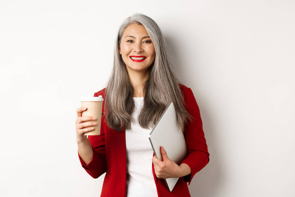 Les affaires. Succès asiatique gestionnaire femme debout avec tasse de café et ordinateur portable, souriant satisfait à la caméra, fond blanc - Photo, image