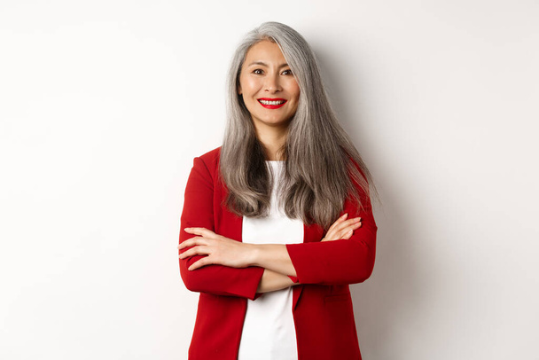 Gente de negocios. Sonriente asiática senior mujer en chaqueta roja, brazos cruzados en el pecho y buscando profesional, de pie sobre fondo blanco - Foto, imagen