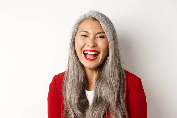 Primer plano de la feliz mujer de negocios asiática con el pelo largo y gris, vistiendo chaqueta roja, riendo y sonriendo alegremente a la cámara, fondo blanco - Foto, imagen