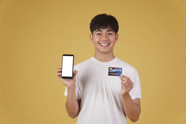 πορτρέτο του χαρούμενος νεαρός ασιάτης ντυμένος άνετα κρατώντας smartphone και πιστωτική κάρτα για ψώνια σε απευθείας σύνδεση απομονωμένο σε κίτρινο φόντο στούντιο - Φωτογραφία, εικόνα