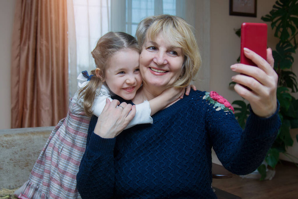 Grootmoeder en kleindochter knuffelen en hebben plezier met het bekijken van een smartphone. Een volwassen vrouw van 50-60 jaar maakt een gezamenlijke selfie foto met haar kleine mooie kleindochter. - Foto, afbeelding