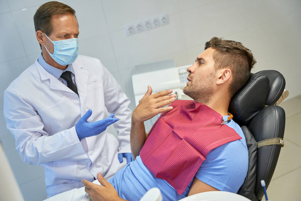Especialista dental problemático explicando algo a su paciente masculino - Foto, Imagen