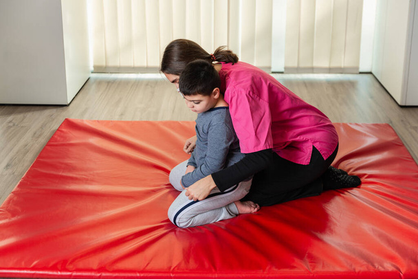 niño discapacitado y fisioterapeuta en una alfombra de gimnasia roja haciendo ejercicios. protección máscara pandémica - Foto, Imagen