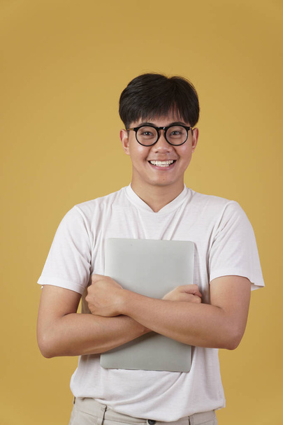 щасливий веселий молодий азіатський чоловік носить студента, одягнений випадково в окуляри, тримаючи ноутбук комп'ютера ізольований на жовтому студійному фоні
 - Фото, зображення