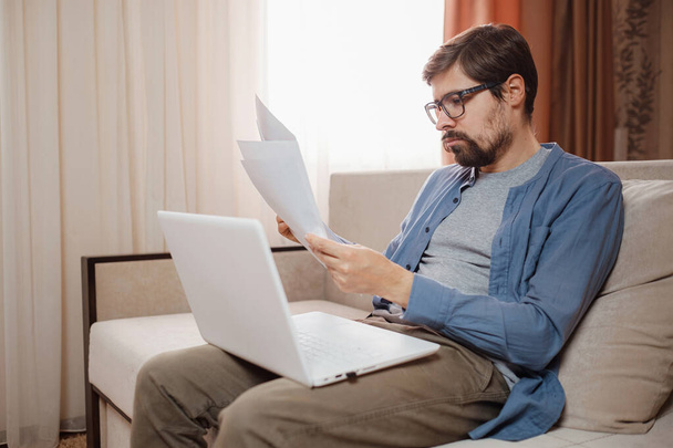 Підприємець в окулярах працює з ноутбуком і зберігає документ в домашньому офісі. Чоловік тримає паперові документи, спілкується онлайн з клієнтами на ноутбуці на робочому місці
. - Фото, зображення