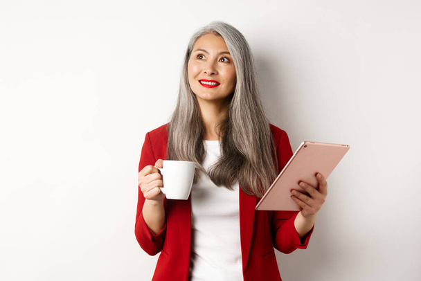 Концепция деловых людей. Азиатская деловая женщина пьет кофе и держит цифровой планшет, улыбаясь вдумчиво в верхнем левом углу, белый фон - Фото, изображение