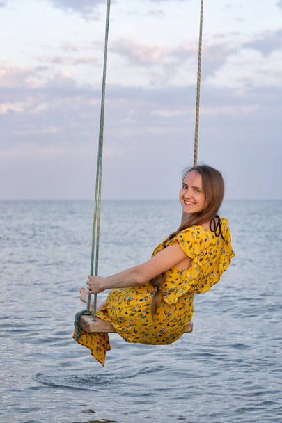 Młoda piękna dziewczyna siedzi na huśtawce liny na tle morza. Rama pionowa - Zdjęcie, obraz