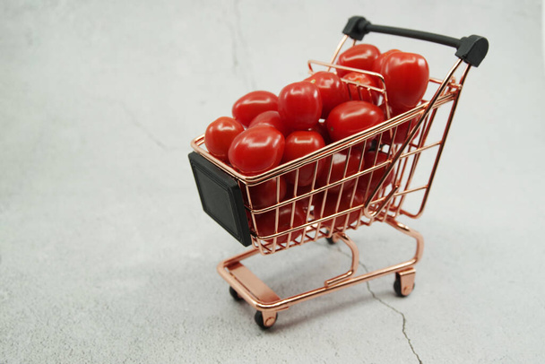 Mini wózek z czerwonymi pomidorami wiśniowymi na szarym tle. Zakupy i żywność kupuje metaforę pojęcia. - Zdjęcie, obraz