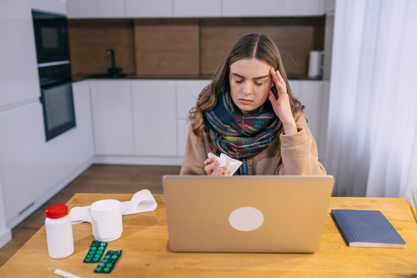 Eine junge kaukasische Frau in Hemd und Schal um den Hals, müde von Arbeit und Krankheit, hält eine Serviette, zu Hause sitzt ein Computer an einem Tisch mit frisch gepresstem Saft.. - Foto, Bild